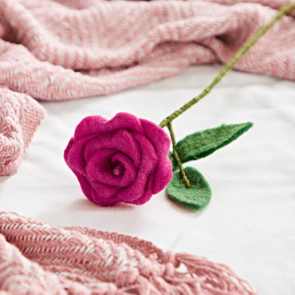 The Felt Rose, in magenta with a green stem and leaf, rests on a white surface beside a pink woven blanket, adding cosy charm to the scene.
