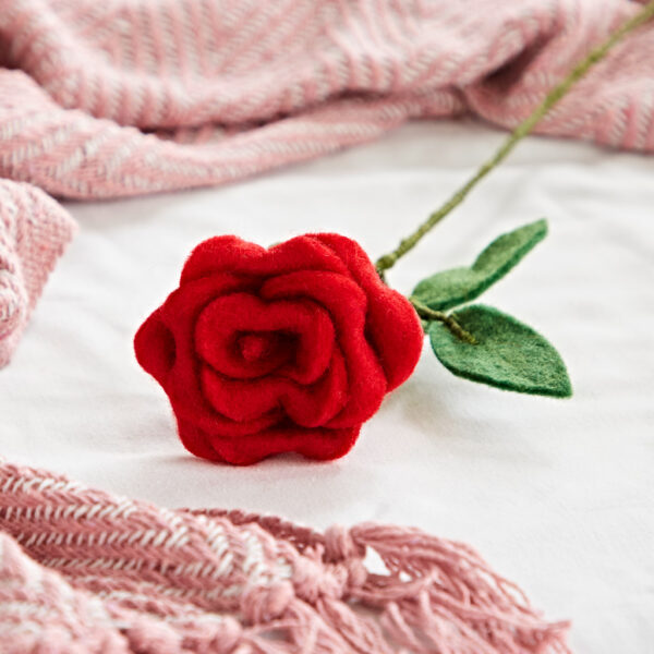 The Felt Rose features vibrant red petals and green felt leaves, displayed on a white surface next to pink textured fabric with fringed edges.