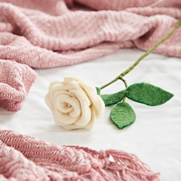 The Felt Rose, featuring white petals and green felt leaves, rests on a white surface beside a pink patterned blanket, bringing delicate charm to the scene.