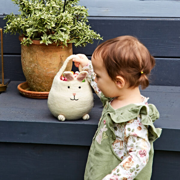 A young child reaches into a Felt Easter Mini Bunny Basket set on a blue wooden step beside a potted plant.