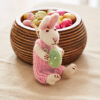 The Felt Rabbit with Egg Hanging Decoration in a pink outfit sits holding a green egg in front of a wooden bowl filled with colourful sweet eggs.