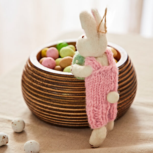 The Felt Rabbit with Egg Hanging Decoration in a pink dress stands next to a wooden bowl of colourful sweet eggs on a beige tablecloth.