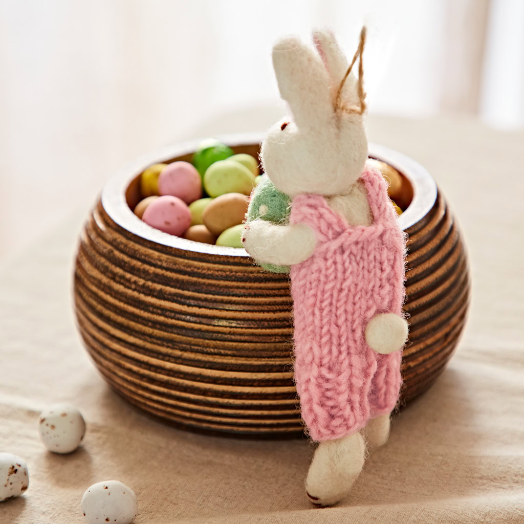 The Felt Rabbit with Egg Hanging Decoration in a pink dress stands next to a wooden bowl of colourful sweet eggs on a beige tablecloth.