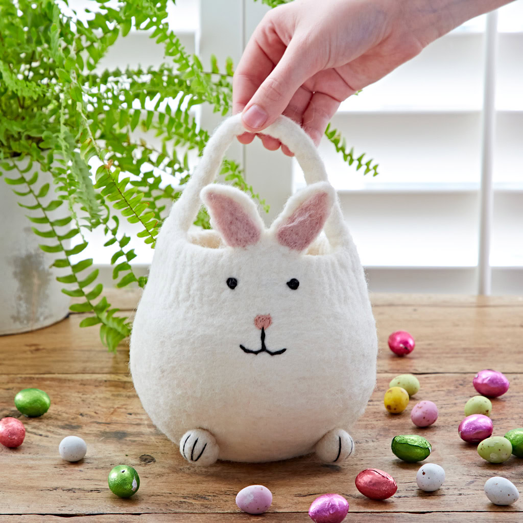 A hand holds the Felt Easter Bunny Basket, shaped like a bunny, on a wooden table with colourful foil-wrapped chocolate eggs and a potted fern in the background.