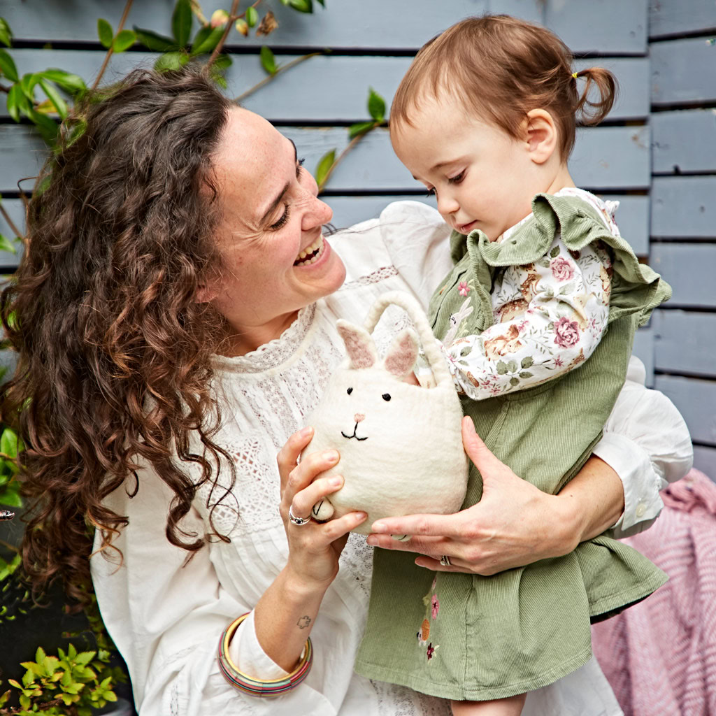 An adult and a young child smile outdoors as the adult holds a small rabbit soft toy next to the colourful Felt Easter Bunny Basket.