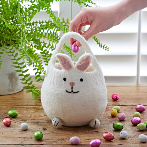 A hand places a pink chocolate egg into the Felt Easter Bunny Basket on a wooden table, with colourful eggs and a fern plant in the background.