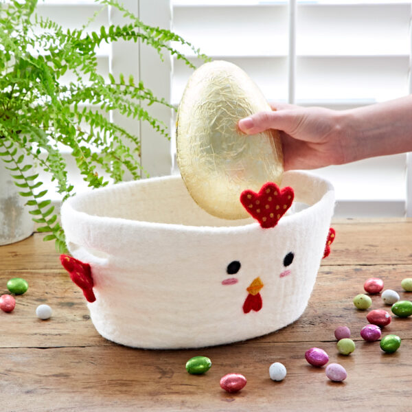 A hand places a large gold-wrapped egg into the Felt Chicken Easter Basket, with colourful foil eggs and a green fern on the nearby wooden table.