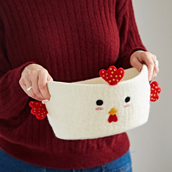 A person holds the Felt Chicken Storage Basket, made of white felt with delightful red and yellow accents on its beak, comb, and wings.