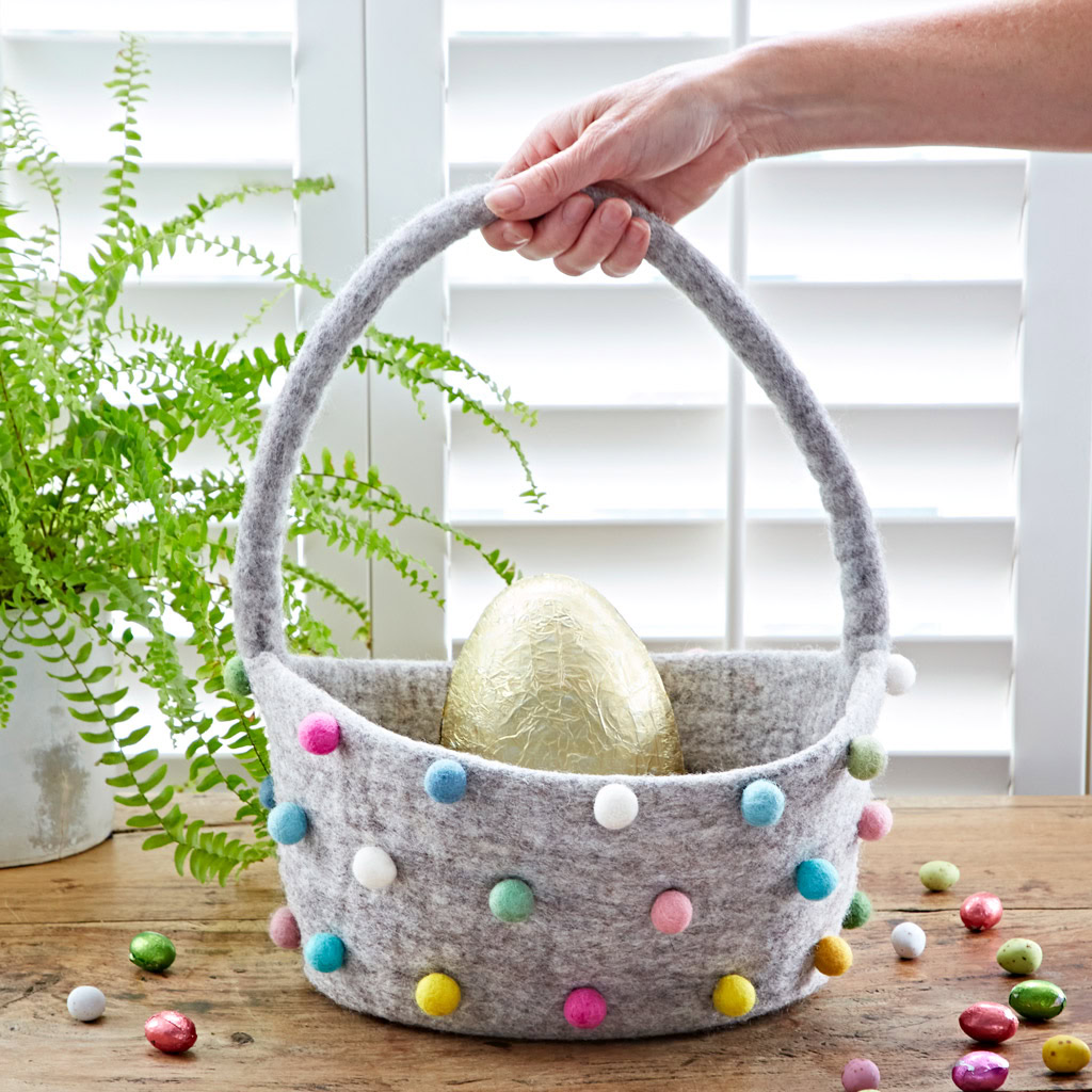 A hand holds a Felt Pom Pom Storage Basket with Handle containing a large gold-wrapped egg, on a wooden table scattered with sweets and set against a potted fern in the background.