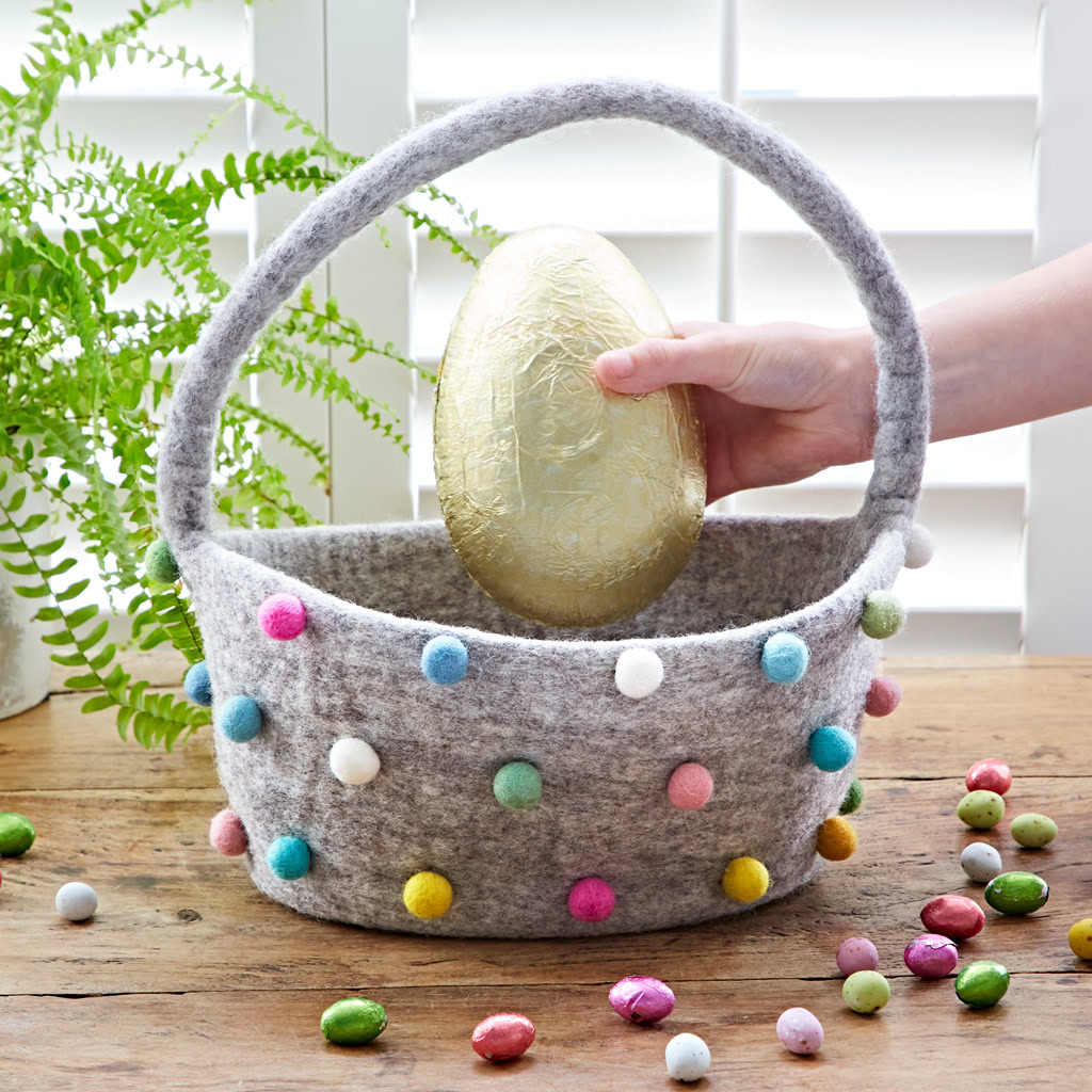 A hand places a large gold-wrapped egg into a Felt Pom Pom Storage Basket with Handle, whilst small eggs are scattered across the wooden table.