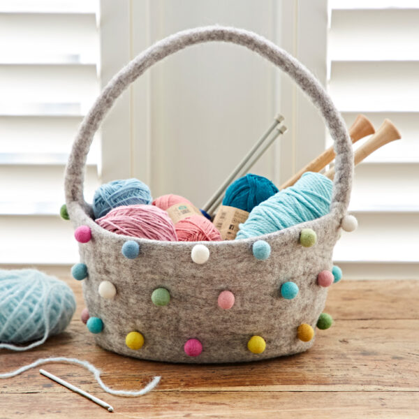 The Felt Pom Pom Storage Basket with Handle organises yarn, needles, and crochet hooks on a wooden table by window shutters, its colourful pom-poms adding a cheerful touch.