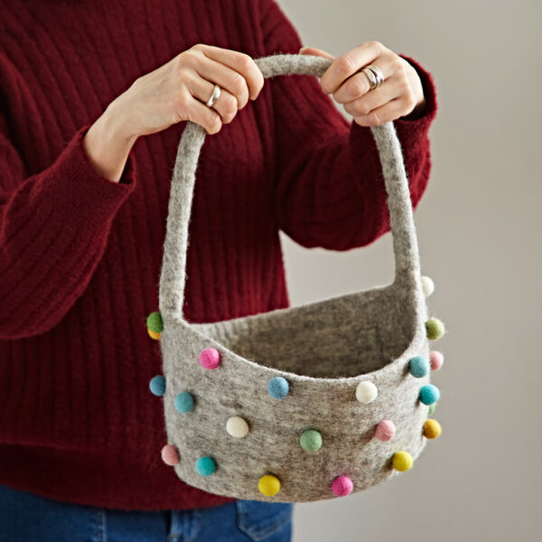 A person in a red jumper holds the Felt Pom Pom Storage Basket with Handle, which features bright multicoloured pom-poms.