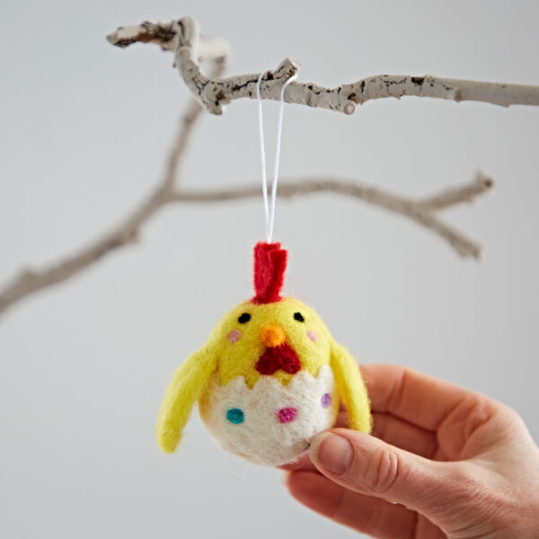 A hand holds the Felt Chick in Egg Hanging Decoration, featuring a yellow chick with a red comb emerging from an egg, suspended from a white branch.