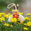 The Felt Bee Hanging Decoration is displayed among yellow wildflowers outdoors, holding an orange flower.
