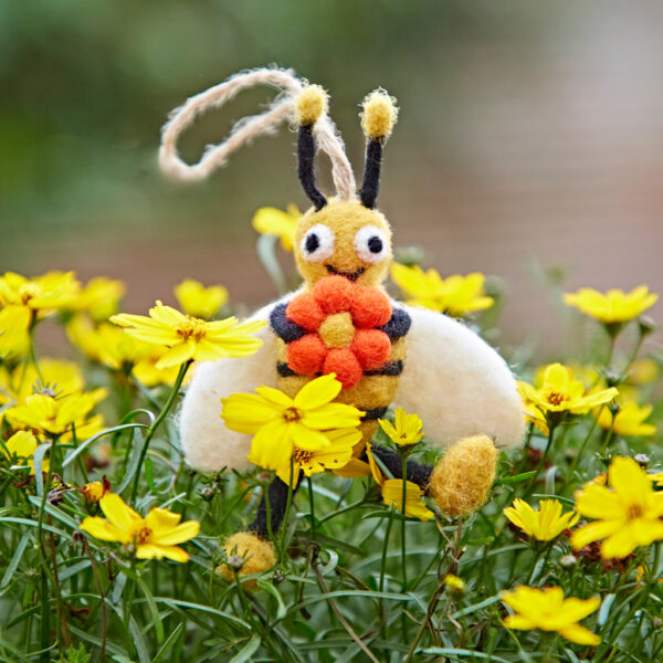 The Felt Bee Hanging Decoration is displayed among yellow wildflowers outdoors, holding an orange flower.