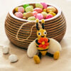 A striped wooden bowl filled with colourful candy eggs sits on a beige surface, with the Felt Bee Hanging Decoration placed in front of the bowl.