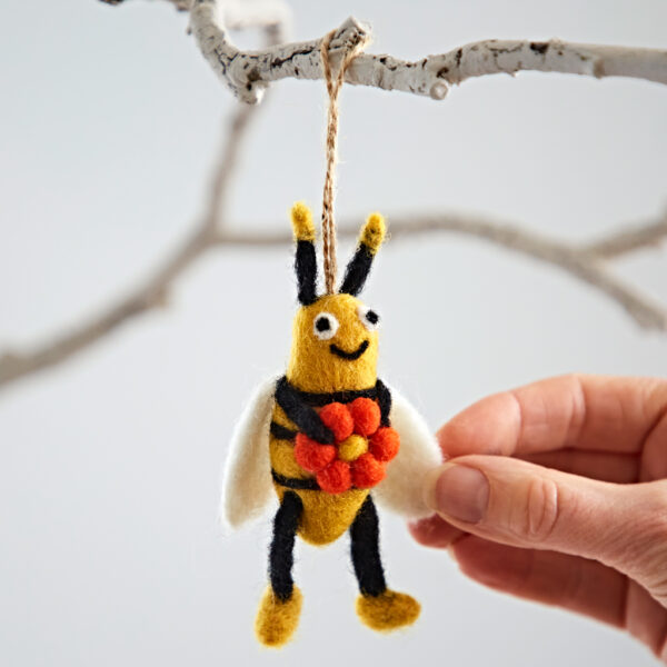 A hand holds the Felt Bee with Flower Hanging Decoration, which features a felt bee with white wings and red and orange pom-pom flowers, all suspended from a white branch.