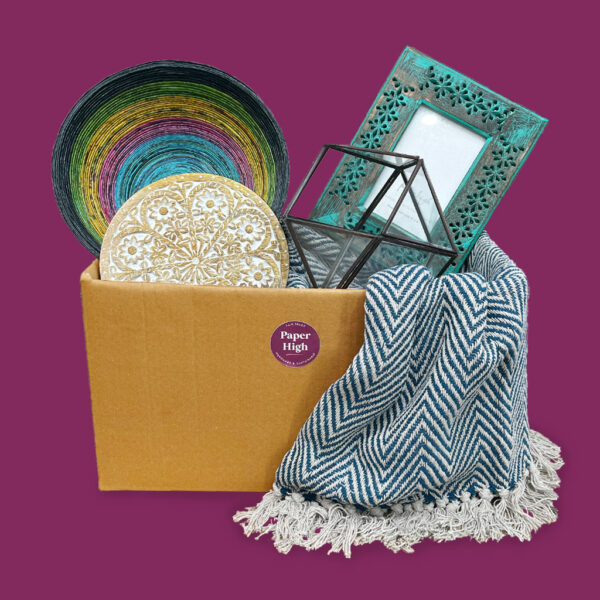 A cardboard box containing an Antique Brass Candle Lantern (Copy), a colourful bowl, decorative coasters, a teal photo frame, and a blue patterned blanket set against a purple background.