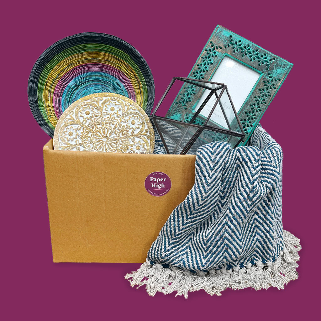 A cardboard box containing an Antique Brass Candle Lantern (Copy), a colourful bowl, decorative coasters, a teal photo frame, and a blue patterned blanket set against a purple background.