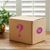The Fair Trade Mystery Box, sealed with a purple question mark and a round "Paper High" label, rests on a wooden table beside a green plant and white window shutters.