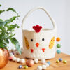 The Felt Easter Chicken Basket with Colourful Ball Charms, shaped like a white chicken, rests on a wooden table amid colourful eggs, pom-poms, and a potted plant.