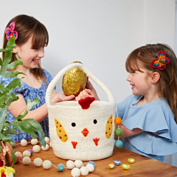 Two young girls smile as they hold a large foil-wrapped egg above a Felt Easter Chicken Basket with Colourful Ball Charms, surrounded by festive decorations and colourful eggs on the table.
