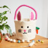 A Felt Easter Chicken Basket with Colourful Ball Charms sits on a wooden table next to Easter eggs, white felt balls, and a potted plant.