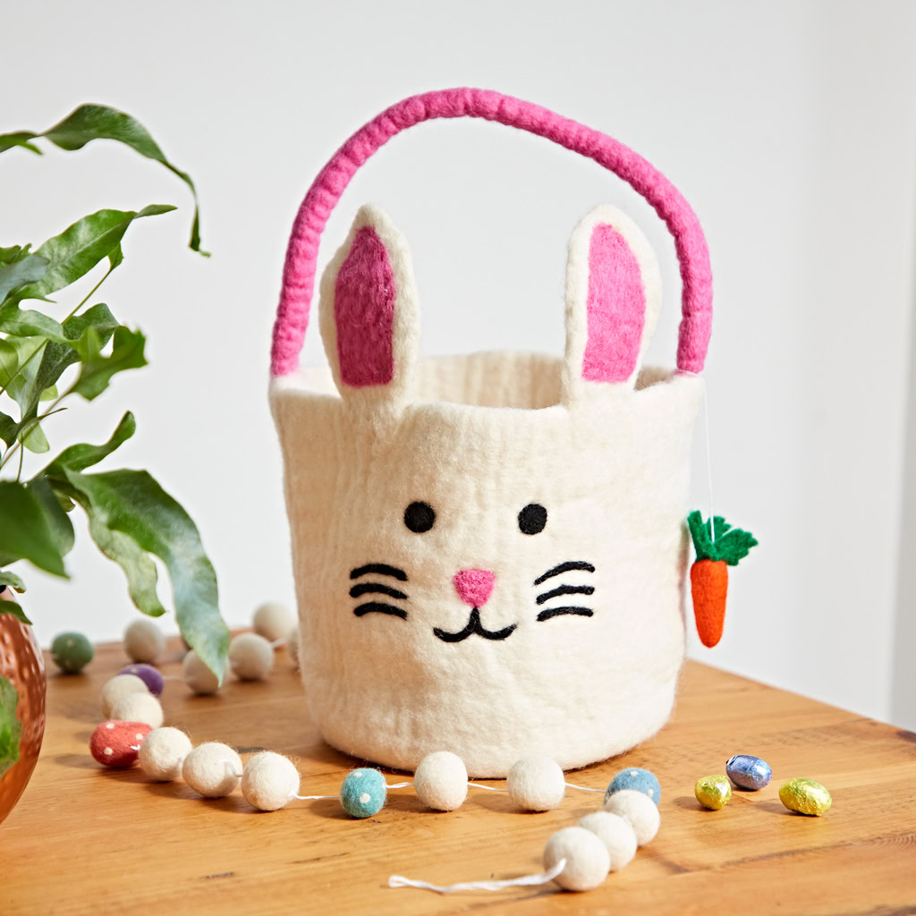 A Felt Easter Chicken Basket with Colourful Ball Charms sits on a wooden table next to Easter eggs, white felt balls, and a potted plant.