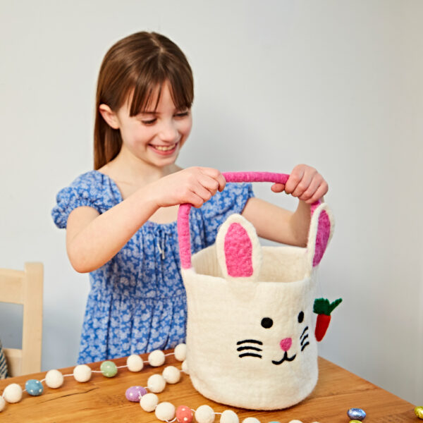 A young girl in a blue dress smiles whilst holding a Felt Easter Chicken Basket with Colourful Ball Charms on a wooden table decorated with eggs and pom-poms.