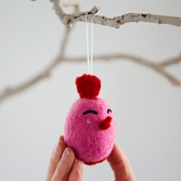 A hand holds the Felt Easter Chick Hanging Decoration—pink with a red crest, beak, and embroidered face—hanging from a white branch.