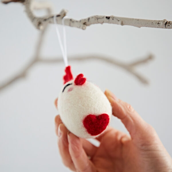 A hand hangs the Felt Easter Chick Hanging Decoration, a white felt ornament with red accents shaped like a chick, from a bare branch.