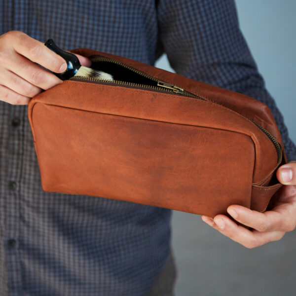 A person in a chequered shirt holds a Reworked Personalised Large Brown Leather Wash Bag, partially unzipped, whilst placing a black item inside.