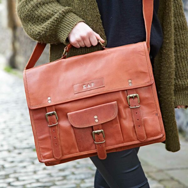Person holding a Personalised Brown Leather Vintage Satchel (Copy) with buckle fastenings and front pocket, standing on a cobbled path.