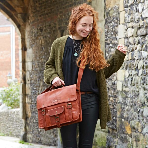 A woman with long red hair, dressed in a dark top, green cardigan, and black trousers, carries the Personalised Brown Leather Vintage Satchel as she walks along a stone wall.