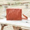 A Personalised Brown Leather Messenger Bag (Copy) with a shoulder strap sits on a wooden picnic table outdoors.