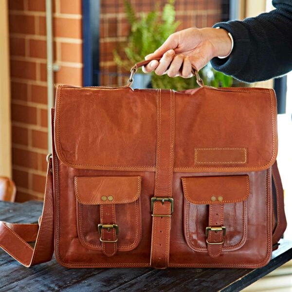 A hand holds the Personalised Brown Leather Satchel Bag (Copy), featuring two front pockets, buckles, a top handle, and a shoulder strap, placed on a dark table indoors.