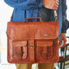 A person holds a Personalised Brown Leather Satchel Bag (Copy) featuring two front pockets, buckles, a handle, and monogrammed with the letters "C.S.H.