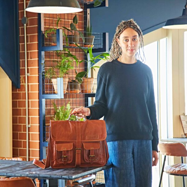A person with plaited hair stands indoors, resting one hand on the Personalised Brown Leather Satchel Bag (Copy) placed on a round table; plants and shelves appear in the background.