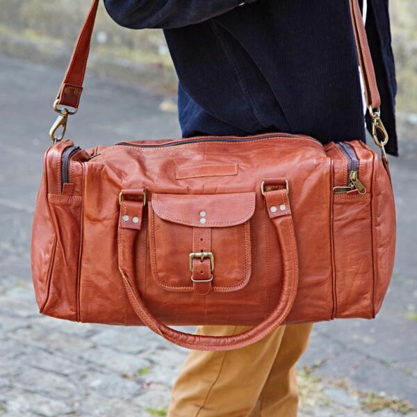 A person stands on a paved surface carrying the Personalised Brown Leather Holdall with Zipped Pockets (Copy), featuring handles and a shoulder strap.