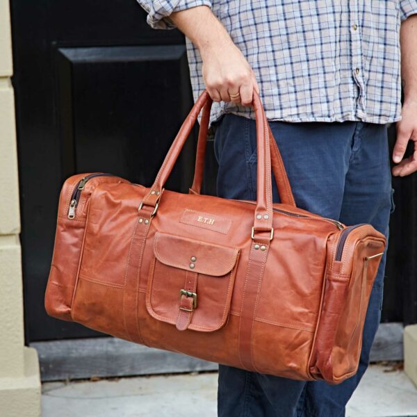 A person in a checked shirt and blue trousers holds the Personalised Brown Leather Holdall with Zipped Pockets, featuring multiple compartments and sturdy handles.