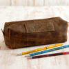 A Reworked Personalised Buffalo Leather Square Pencil Case with a zip rests on a light wooden surface beside three coloured pencils.