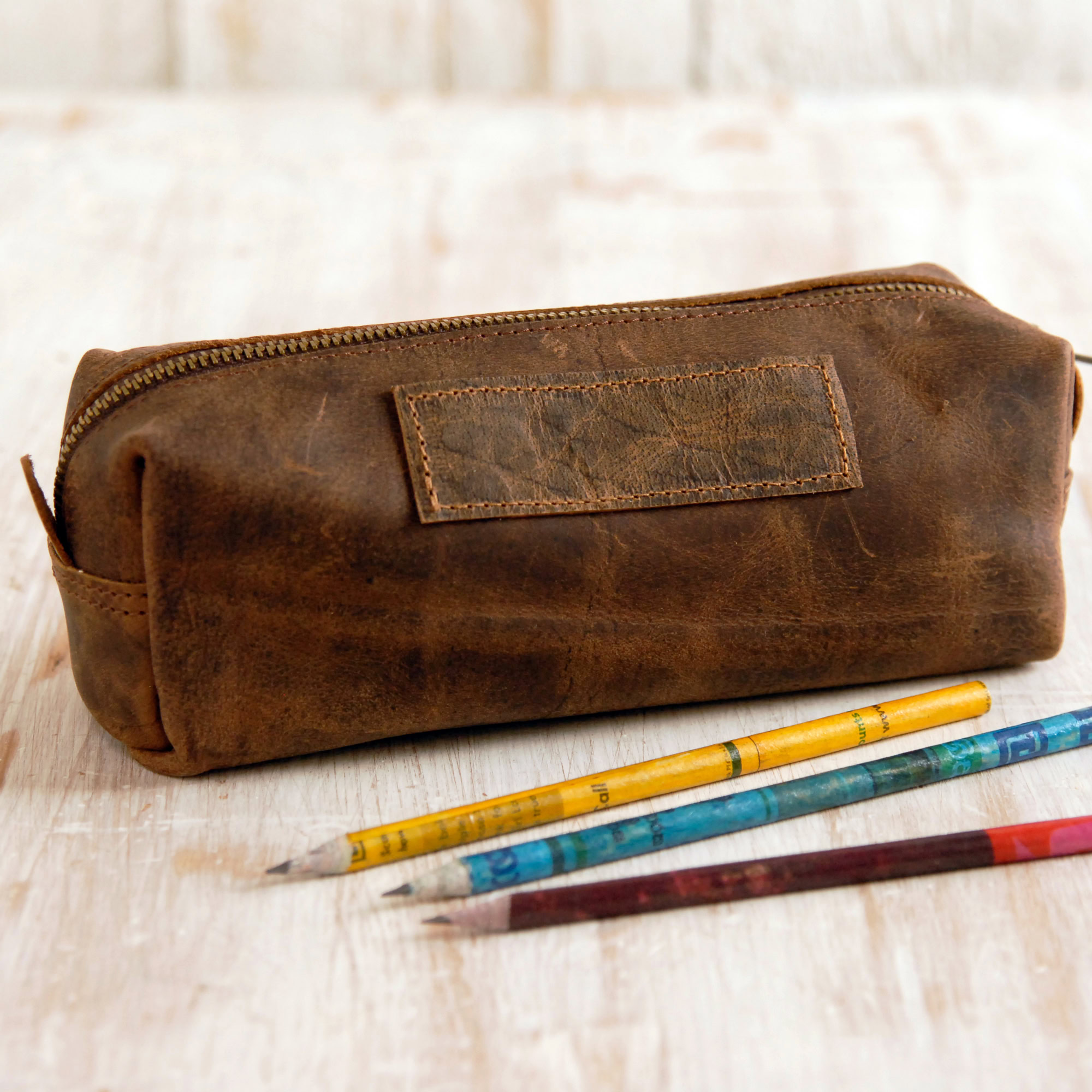 A Reworked Personalised Buffalo Leather Square Pencil Case with a zip rests on a light wooden surface beside three coloured pencils.