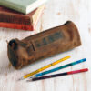 A Reworked Personalised Buffalo Leather Round Pencil Case sits beside three colourful pencils and stacked books on a light wooden surface.