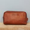 The Personalised Large Brown Leather Wash Bag rests on a wooden surface with a plain light grey wall in the background.