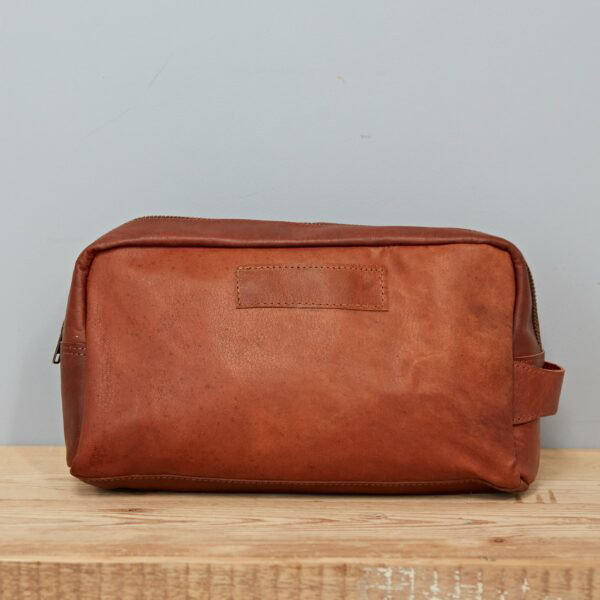 The Personalised Large Brown Leather Wash Bag rests on a wooden surface with a plain light grey wall in the background.