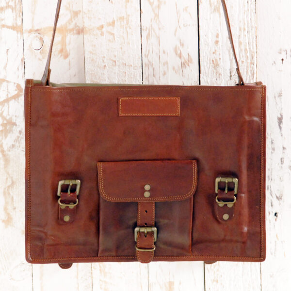 Personalised Brown Leather Vintage Satchel (Copy) featuring a front pocket, metal buckles, and a shoulder strap, displayed against a white wooden background.