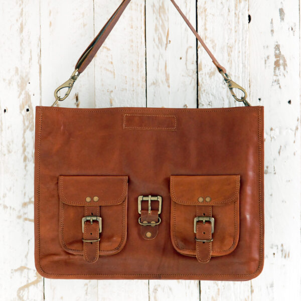 Personalised Brown Leather Satchel Bag (Copy) featuring two front pockets, buckle fastenings, and a shoulder strap, displayed against a white wooden background.