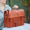 Someone holds a Personalised Brown Leather Vintage Satchel (Copy) with a handle, buckles, front pocket, and "KAF" initials on a label, resting on a white outdoor table.