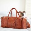 A Personalised Brown Leather Holdall with Zipped Pockets rests on a bed in a bright room with white walls and shutters, as a small dog peeps out from behind it.