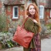 A woman with long red hair, in a green cardigan and checked dress, carries the Personalised Brown Leather Zipped Tote Shopping Bag over her shoulder while standing outside near brick and stone houses.